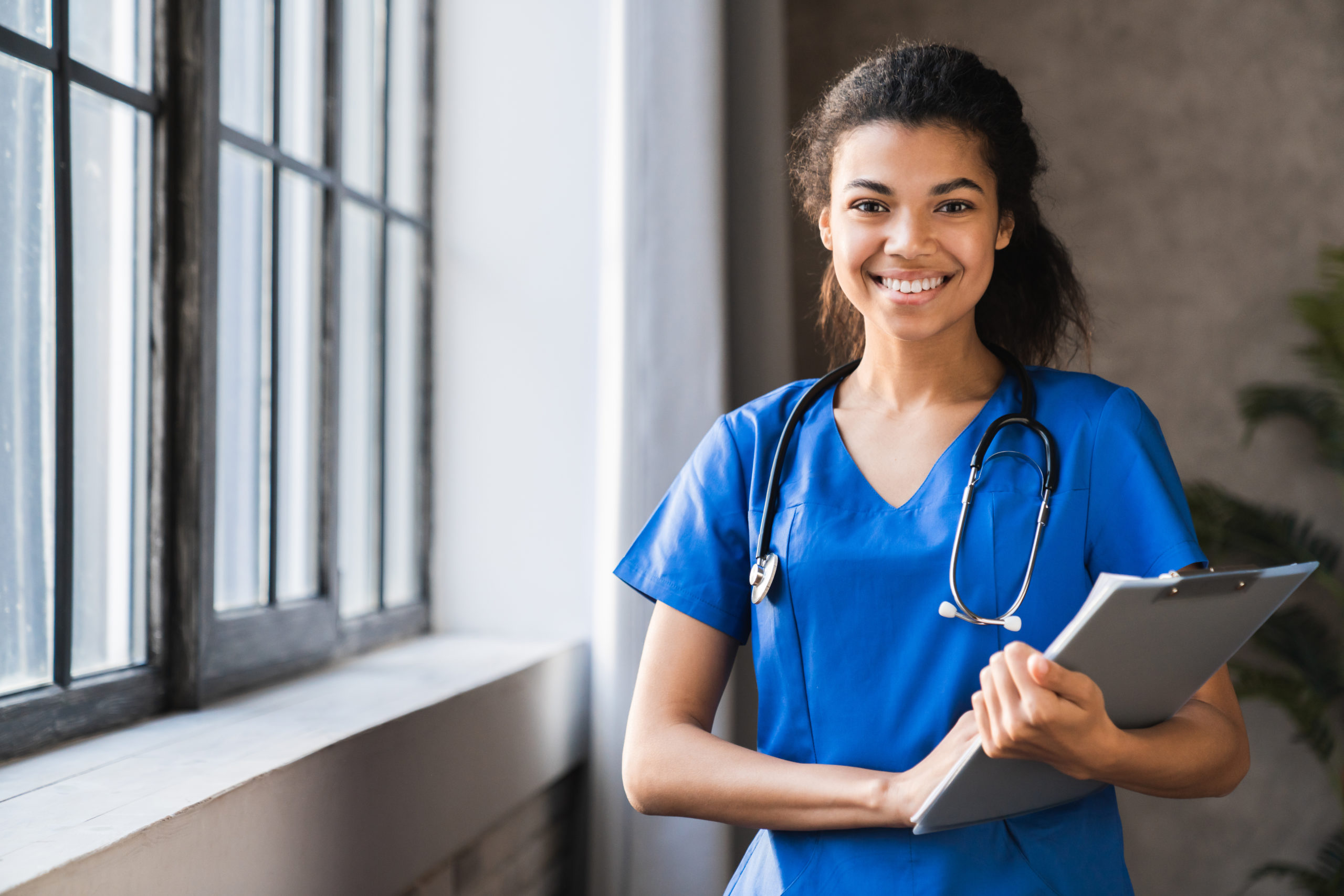 A nursing assistant smiles with a clipboard in hand and stethoscope around her neck.